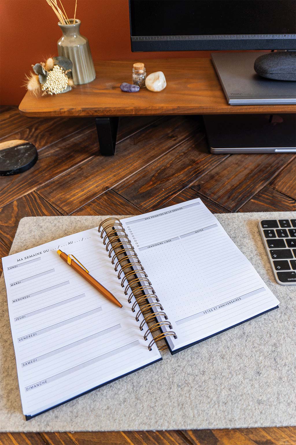 Photo de l'intérieur de l'agenda Jill qui est posé sur un sous-main sur un bureau. On aperçoit également un ordinateur, son écran et son clavier ainsi que des éléments de décorations tels que des fleurs séchées ou des pierres.