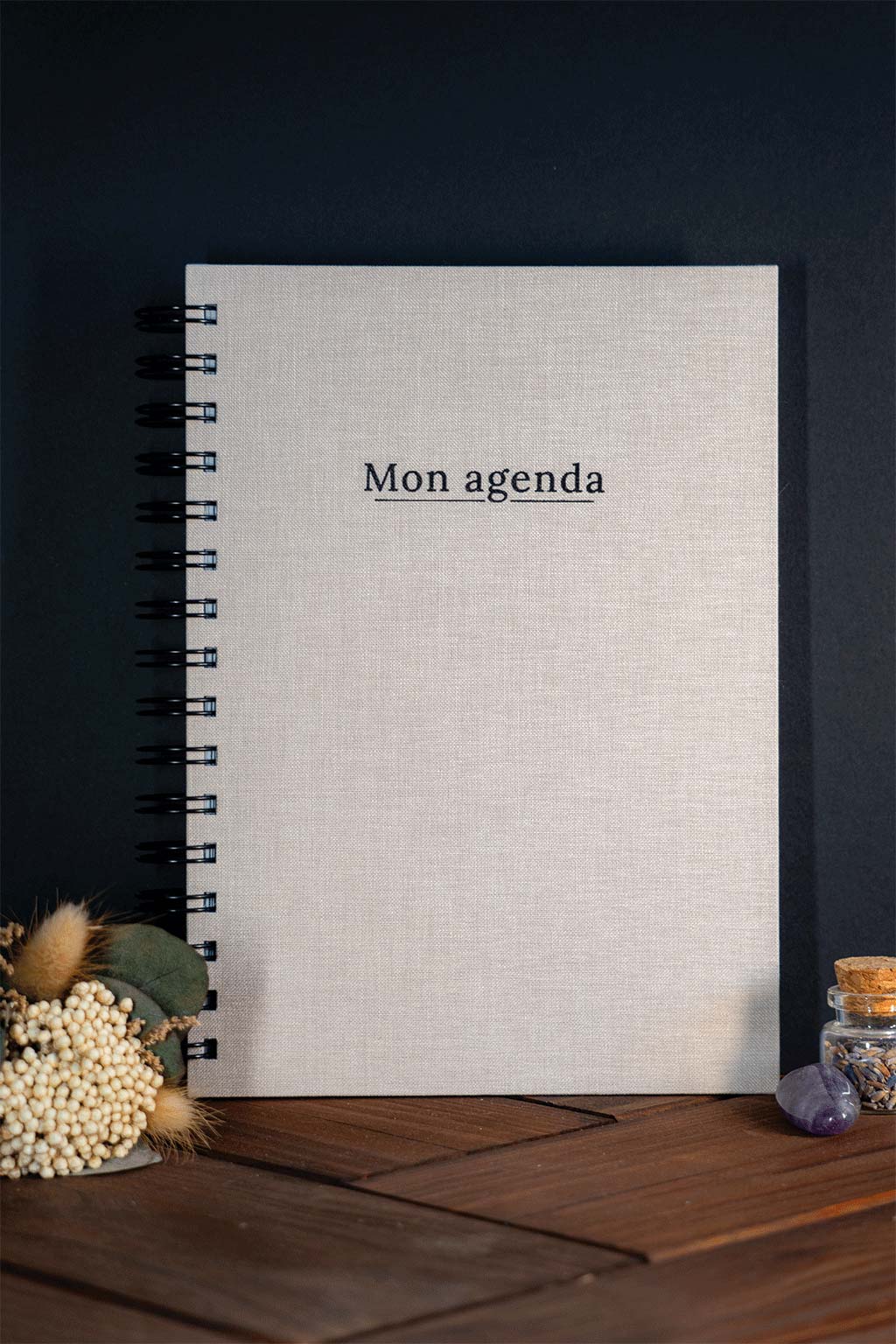 Photo de "Mon agenda" recouvert d'une toile beige avec l'inscription "Mon agenda" et la spirale en noir. Le carnet est posé à la verticale devant un fond noir et on aperçoit également des éléments de décoration tels des fleurs séchés ou une pierre.