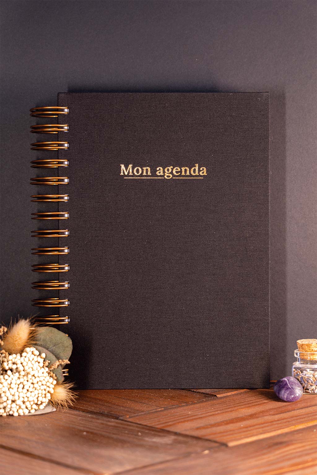 Photo de "Mon agenda" recouvert d'une toile noire avec l'inscription "Mon agenda" et la spirale en couleur bronze. Le carnet est posé à la verticale devant un fond noir et on aperçoit également des éléments de décoration tels des fleurs séchés ou une pierre.
