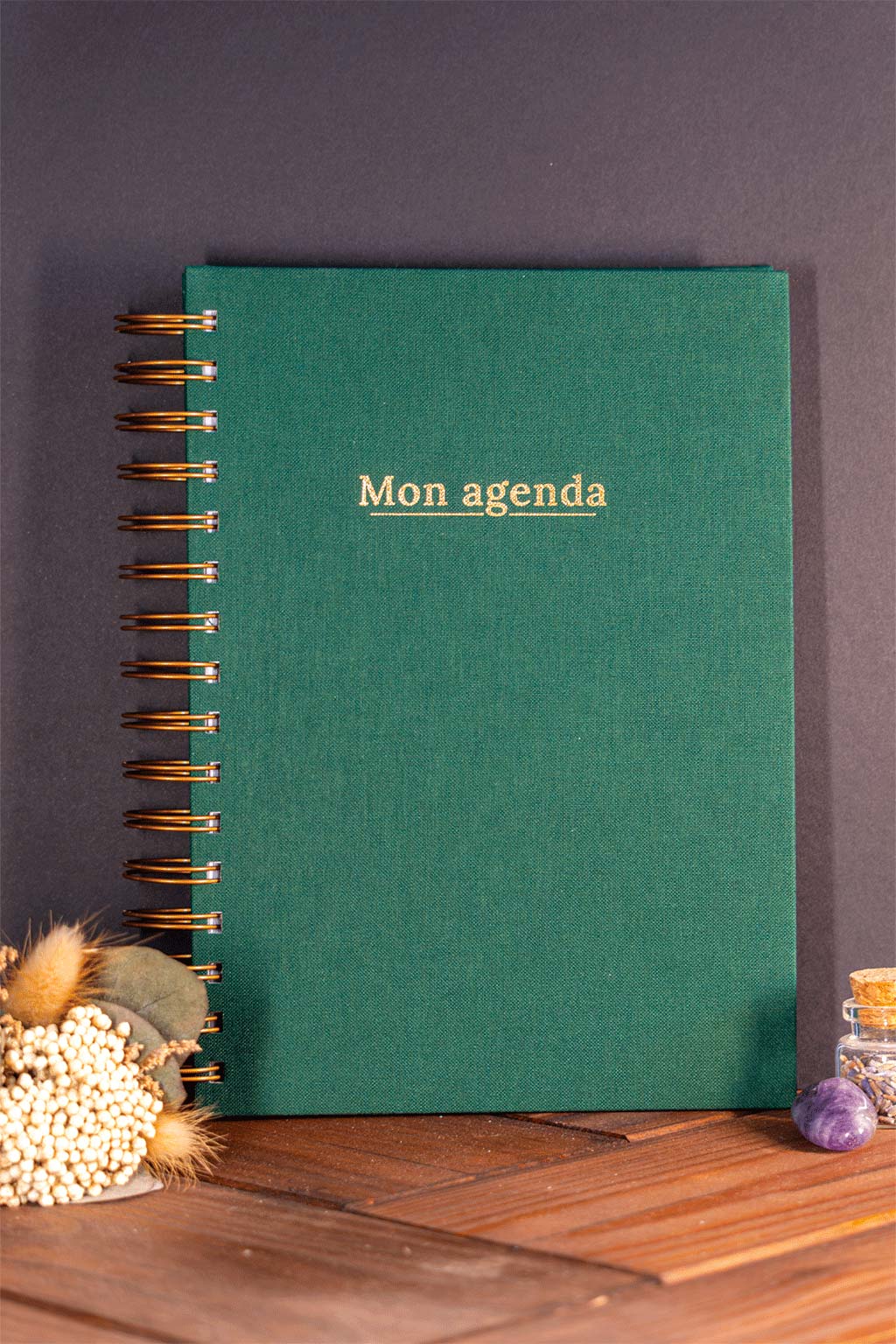 Photo de "Mon agenda" recouvert d'une toile vert sapin avec l'inscription "Mon agenda" et la spirale en couleur bronze. Le carnet est posé à la verticale devant un fond noir et on aperçoit également des éléments de décoration tels des fleurs séchés ou une pierre.