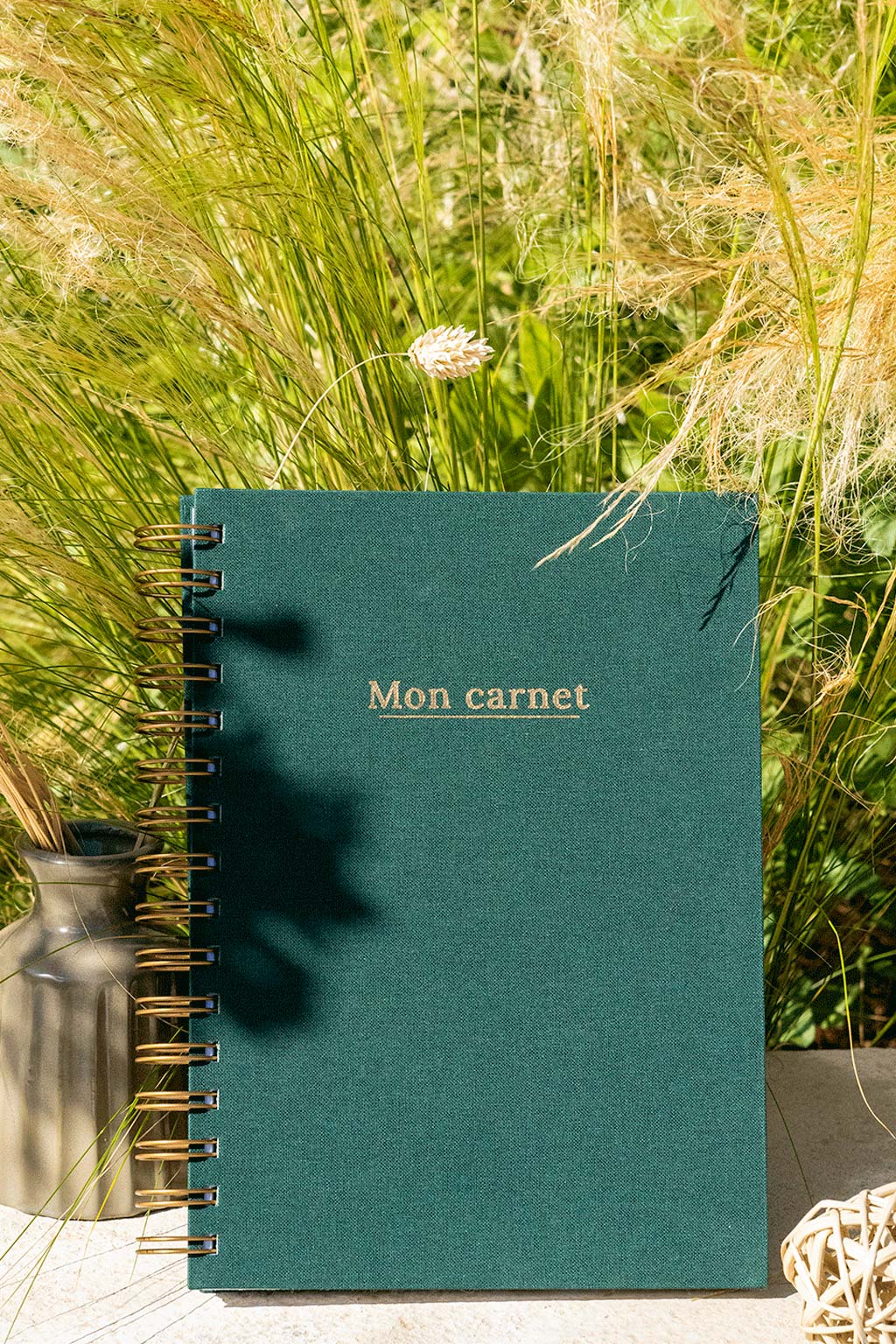 Photo de "Mon Carnet" recouvert d'une toile vert sapin avec l'inscription "Mon carnet" et la spirale de couleur bronze. Le carnet est posé à la verticale devant des graminées et on aperçoit également des éléments de décoration tels qu'un petit vase avec des fleurs et végétaux séchés.