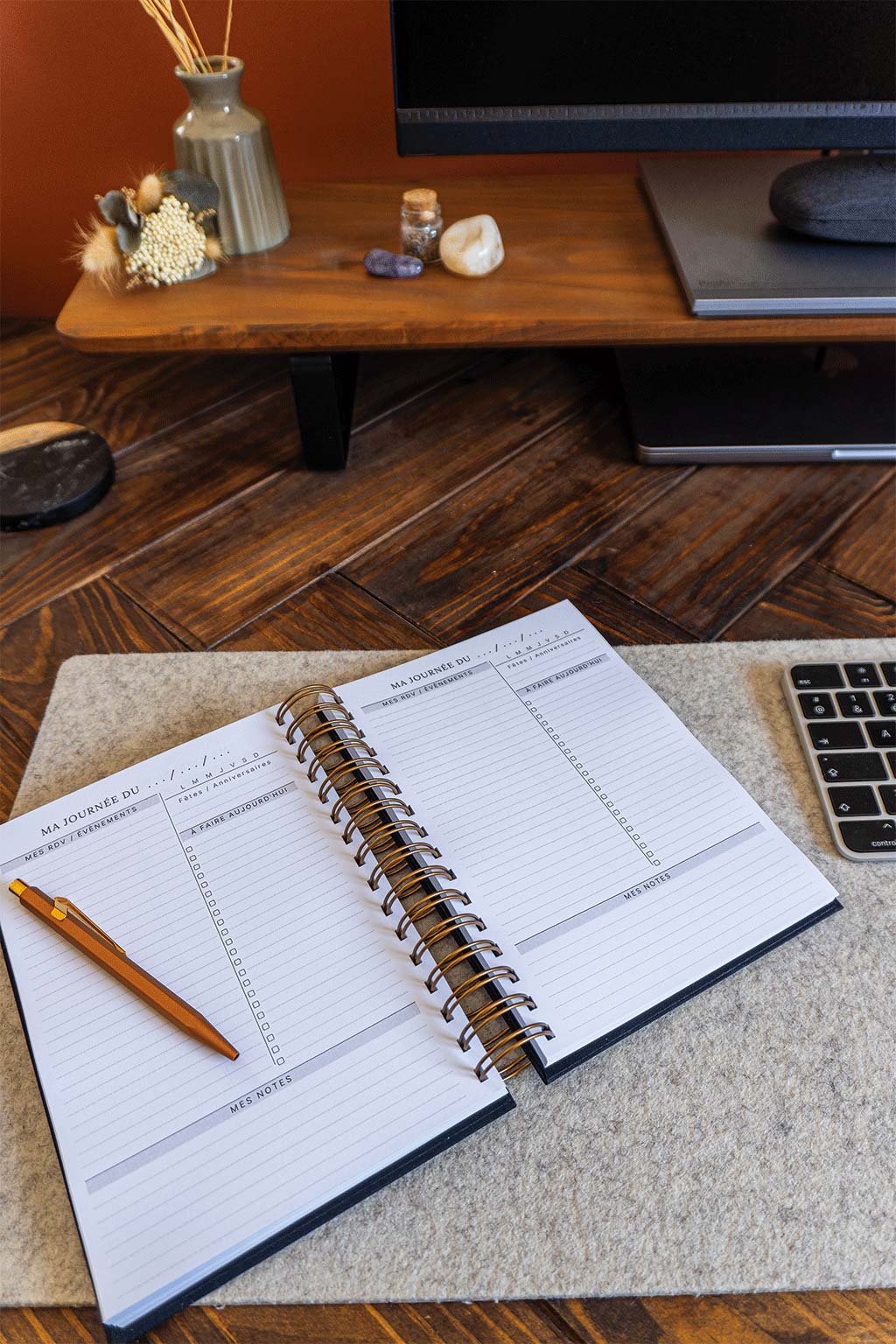 Photo de l'intérieur de l'agenda Alix qui est posé sur un sous-main sur un bureau. On aperçoit également un ordinateur, son écran et son clavier ainsi que des éléments de décorations tels que des fleurs séchées ou des pierres.