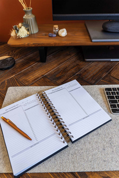 Photo de l'intérieur de l'agenda Alix qui est posé sur un sous-main sur un bureau. On aperçoit également un ordinateur, son écran et son clavier ainsi que des éléments de décorations tels que des fleurs séchées ou des pierres.