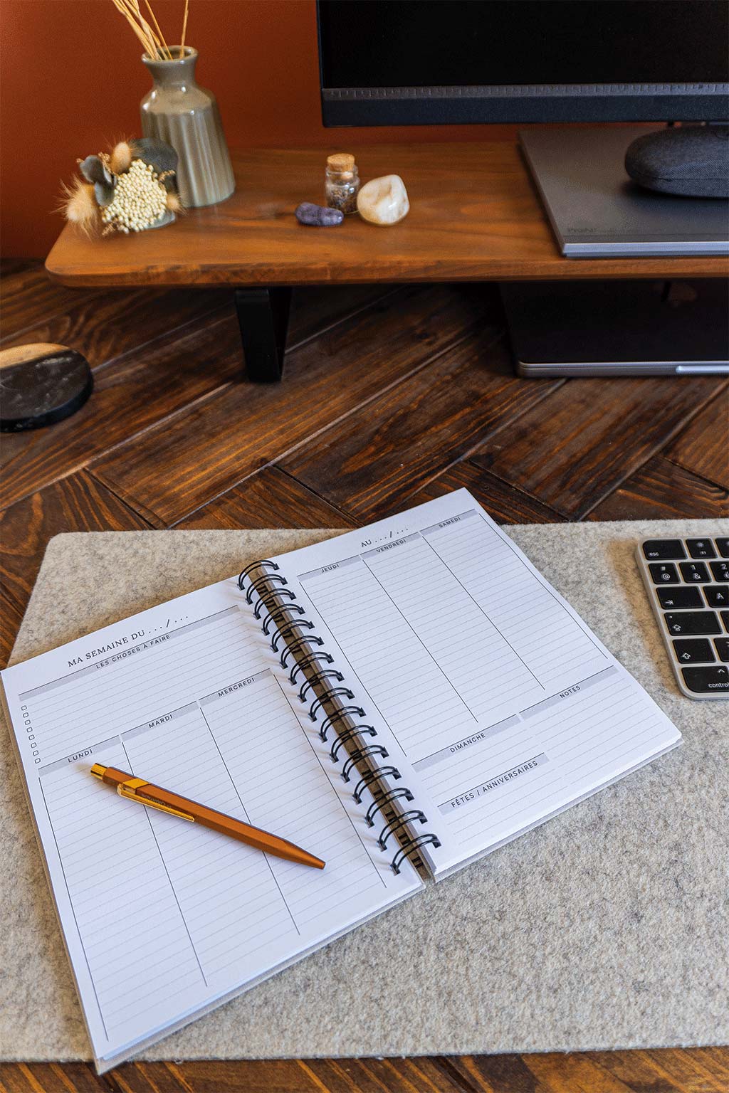 Photo de l'intérieur de l'agenda Gaëlle qui est posé sur un sous-main sur un bureau. On aperçoit également un ordinateur, son écran et son clavier ainsi que des éléments de décorations tels que des fleurs séchées ou des pierres.