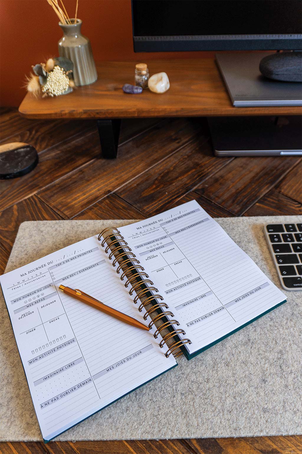 Photo de l'intérieur de l'agenda Lea qui est posé sur un sous-main sur un bureau. On aperçoit également un ordinateur, son écran et son clavier ainsi que des éléments de décorations tels que des fleurs séchées ou des pierres.