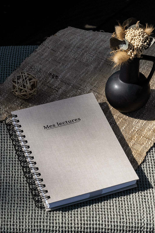 Photo du carnet "Mes lectures" recouvert d'une toile beige avec l'inscription "Mes lectures" et la spirale en noir. Le carnet est posé sur une toile de jute ainsi qu'une serviette couleur kaki, et on observe également quelques éléments de décoration tels  qu'un petit vase contenant des fleurs séchées.