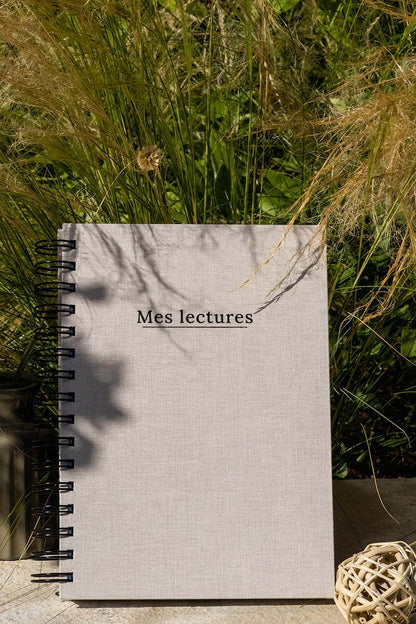 Photo du carnet "Mes lectures" recouvert d'une toile beige avec l'inscription "Mes lectures" et la spirale en noir. Le carnet est posé à la verticale devant des graminées et on aperçoit également des éléments de décoration tels qu'un petit vase avec des fleurs et végétaux séchés.
