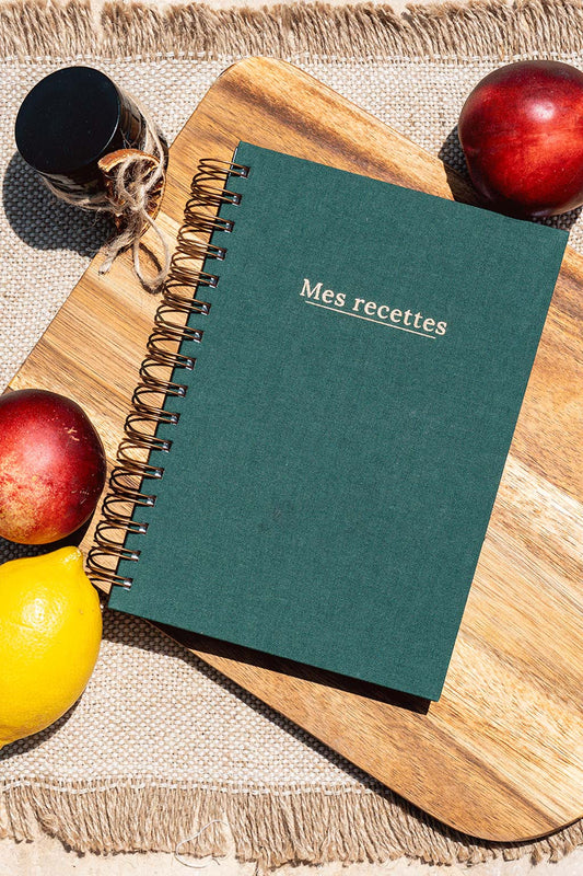 Photo du carnet "Mes recettes" recouvert d'une toile vert sapin avec l'inscription "Mes recettes " et la spirale de couleur bronze. Le carnet est posé sur une planche à découper en bois et entouré de fruits comme des nectarines ou des citrons.