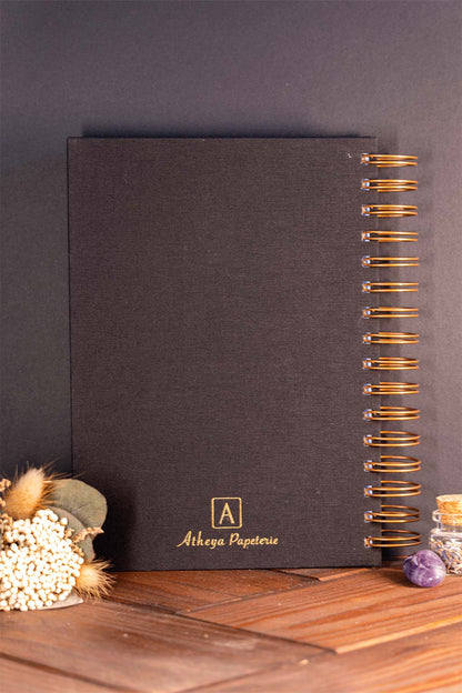 Photo de la quatrième de couverture de "Mon agenda" recouvert d'une toile noire avec l'inscription "Atheya Papeterie" et la spirale en couleur bronze. Le carnet est posé à la verticale devant un fond noir et on aperçoit également des éléments de décoration tels des fleurs séchés ou une pierre.