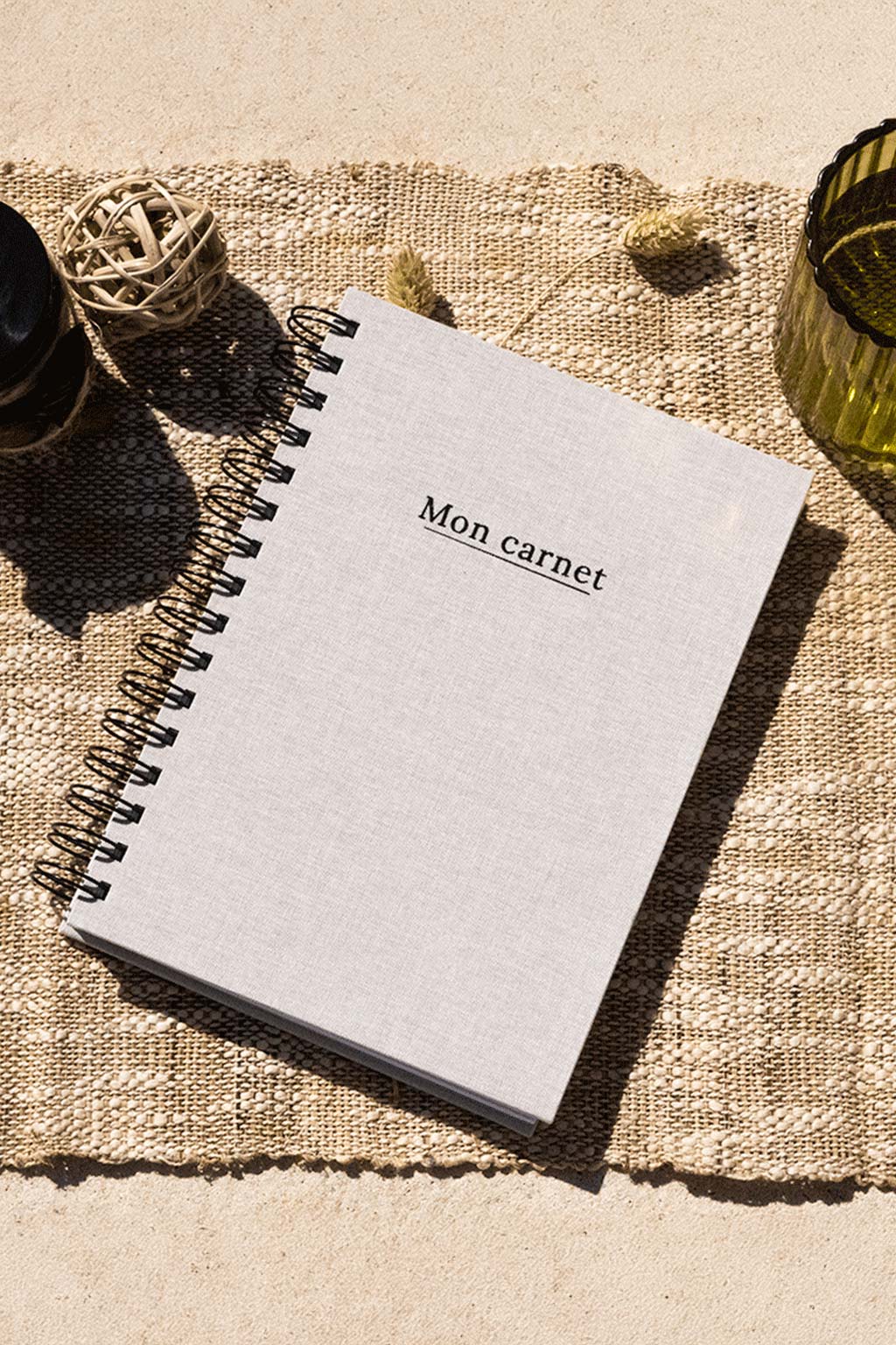 Photo de  "Mon Carnet" recouvert d'une toile beige avec l'inscription "Mon carnet" et la spirale en noir. Le carnet est posé sur une toile de jute et on observe également quelques éléments de décoration comme des fleurs, séchées, un verre ou encore un récipient en verre foncé.