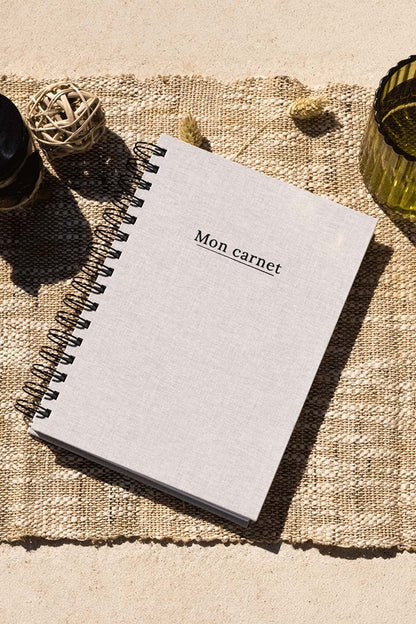 Photo de  "Mon Carnet" recouvert d'une toile beige avec l'inscription "Mon carnet" et la spirale en noir. Le carnet est posé sur une toile de jute et on observe également quelques éléments de décoration comme des fleurs, séchées, un verre ou encore un récipient en verre foncé.