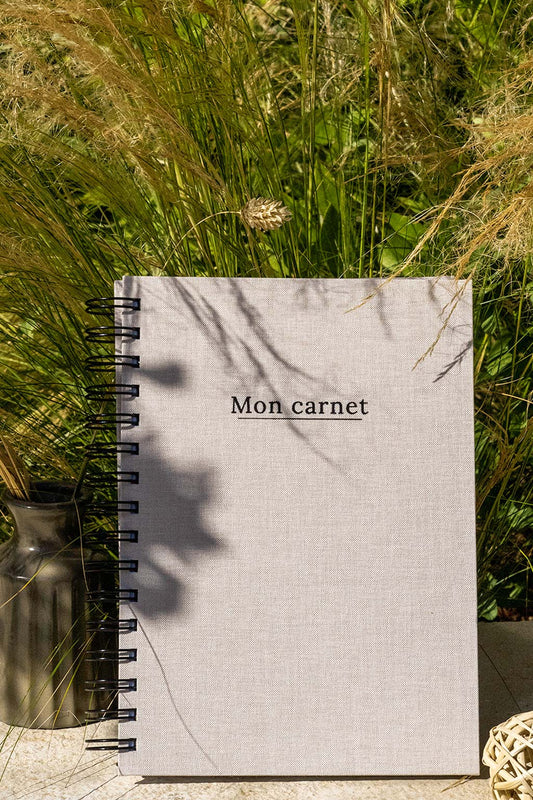 Photo de  "Mon Carnet" recouvert d'une toile beige avec l'inscription "Mon carnet" et la spirale en noir. Le carnet est posé à la verticale devant des graminées et on aperçoit également des éléments de décoration tels qu'un petit vase avec des fleurs et végétaux séchés.