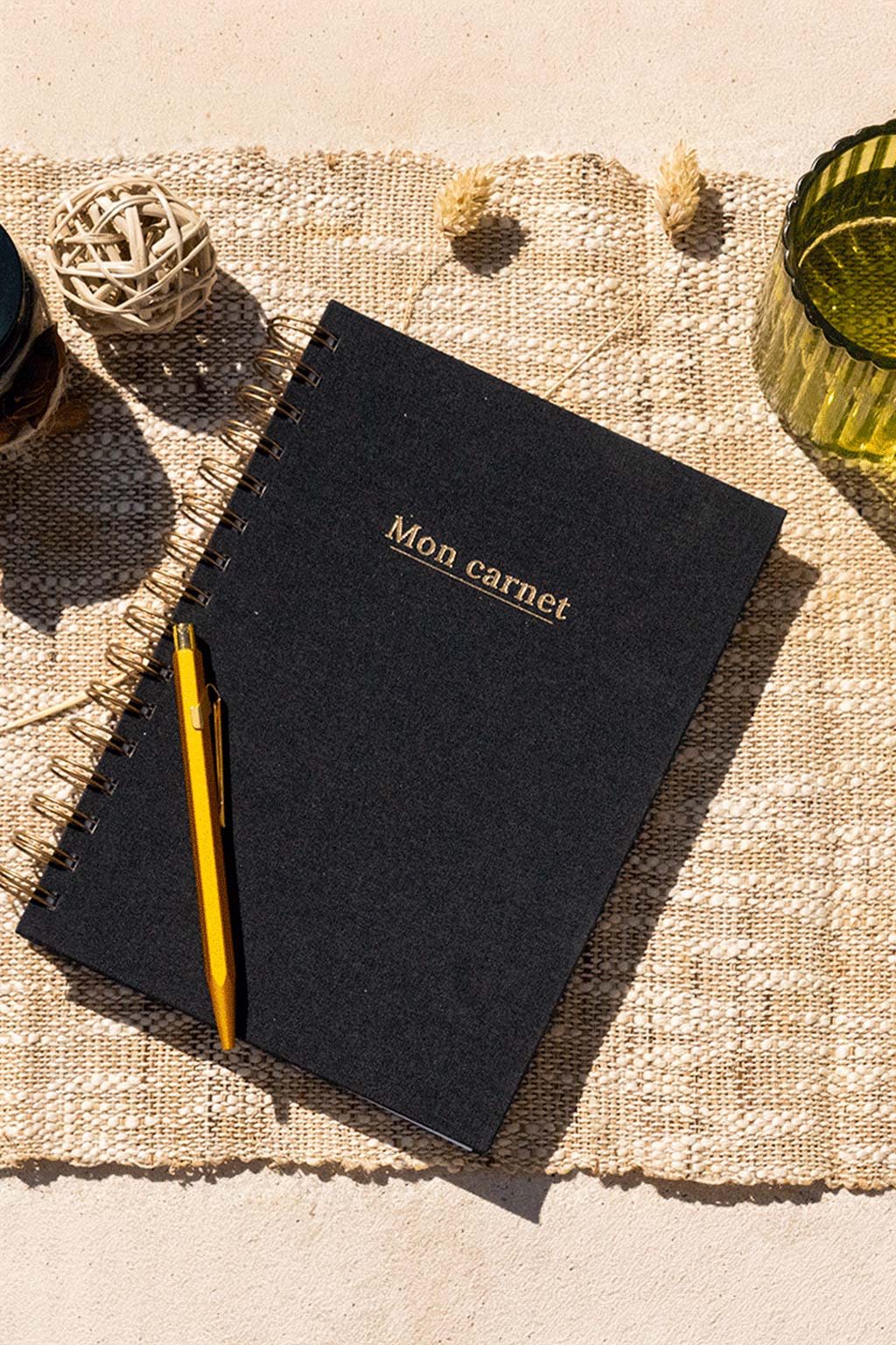 Photo de  "Mon Carnet" recouvert d'une toile noire avec l'inscription "Mon carnet" et la spirale de couleur bronze. Le carnet est posé sur une toile de jute et on observe également quelques éléments de décoration comme des fleurs séchées, un verre ou encore un récipient en verre foncé.