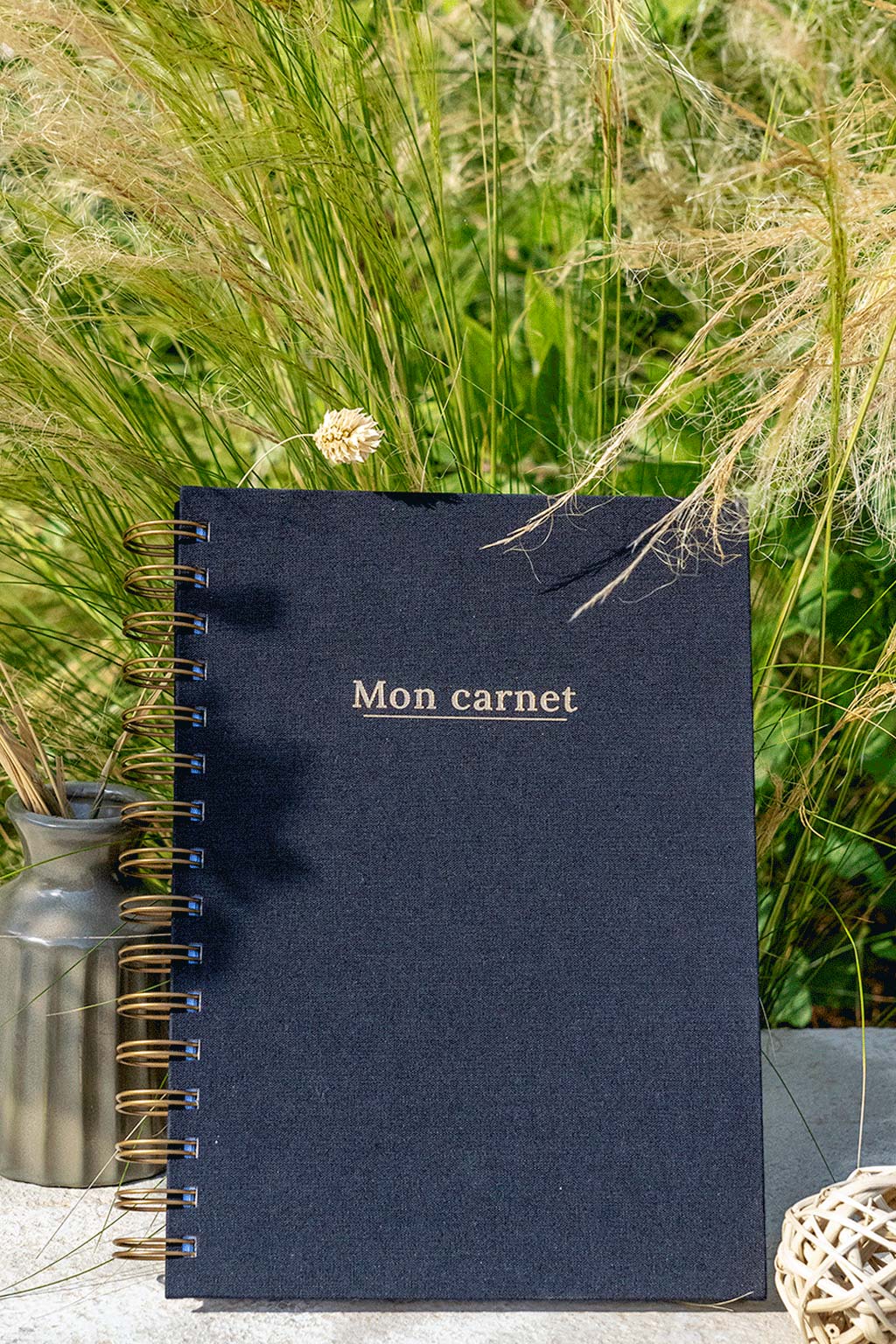 Photo de  "Mon Carnet" recouvert d'une toile noire avec l'inscription "Mon carnet" et la spirale de couleur bronze. Le carnet est posé à la verticale devant des graminées et on aperçoit également des éléments de décoration tels qu'un petit vase avec des fleurs et végétaux séchés.