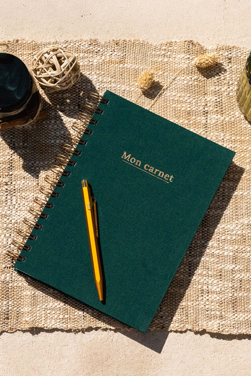 Photo de  "Mon Carnet" recouvert d'une toile vert sapin avec l'inscription "Mon carnet" et la spirale de couleur bronze. Le carnet est posé sur une toile de jute et on observe également quelques éléments de décoration comme des fleurs séchées, un verre ou encore un récipient en verre foncé.