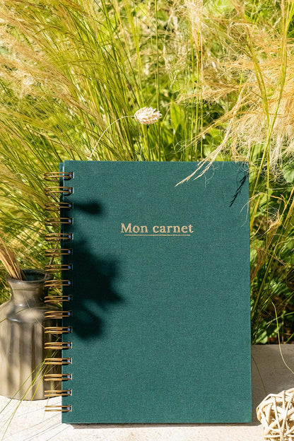 Photo de  "Mon Carnet" recouvert d'une toile vert sapin avec l'inscription "Mon carnet" et la spirale de couleur bronze. Le carnet est posé à la verticale devant des graminées et on aperçoit également des éléments de décoration tels qu'un petit vase avec des fleurs et végétaux séchés.